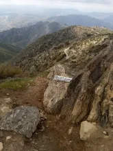 Rocky mountain trail with "Mount Lukens" sign, overlooking distant hills and valley.